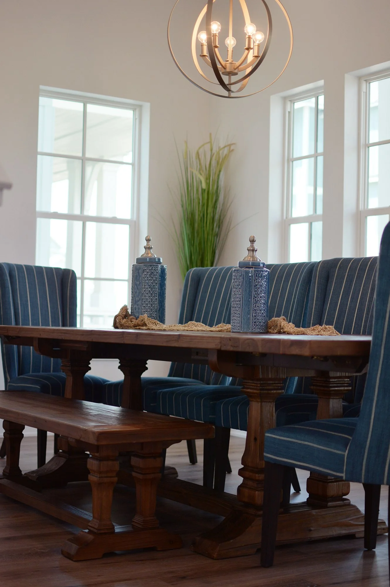 A dining room featuring a wooden table with a textured table runner, decorated with two ornate blue vases with metallic lids, and surrounded by upholstered chairs with blue and white striped fabric. A large window provides natural light, and a modern chandelier hangs from the ceiling. In the background, a tall green plant is placed against a white wall.