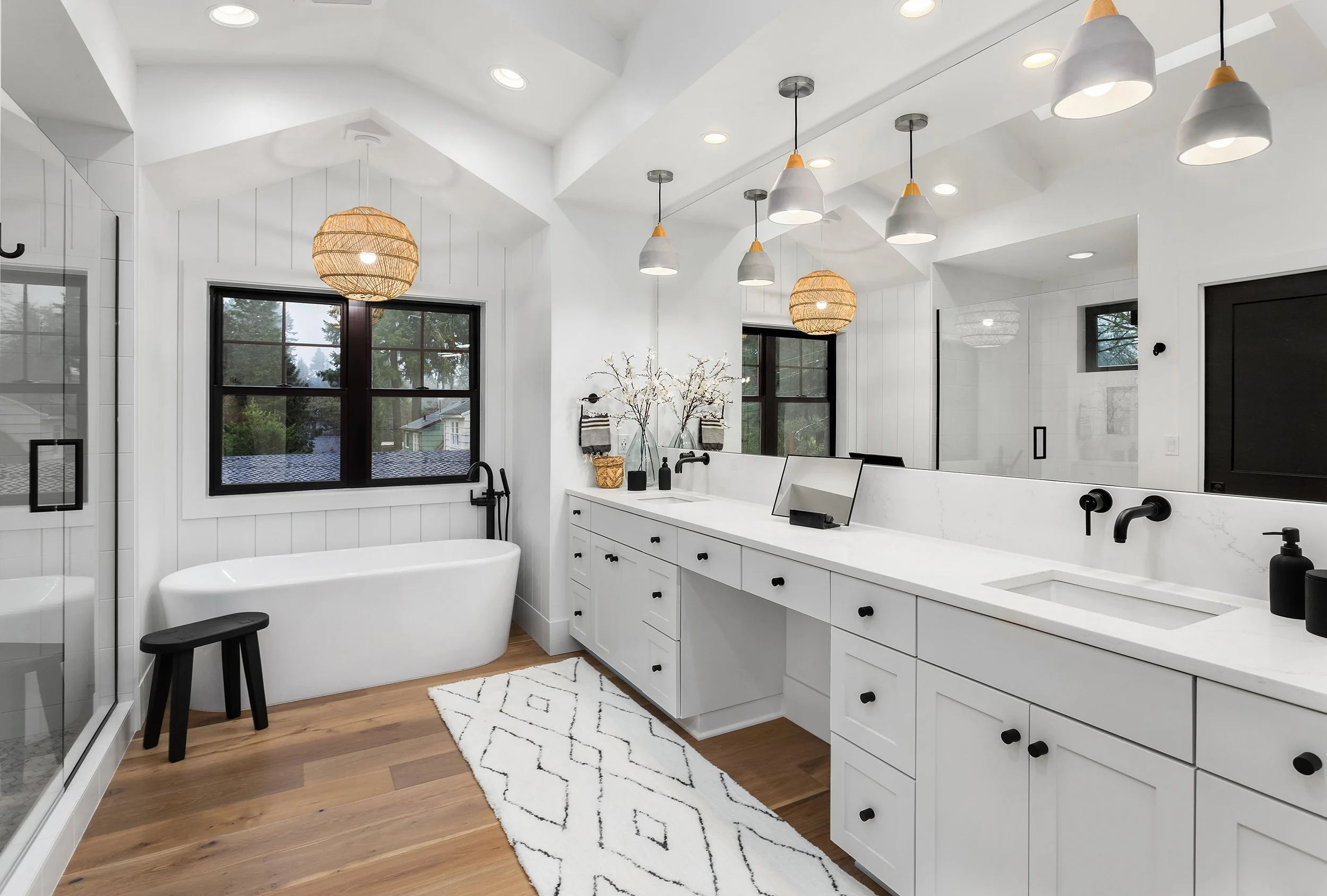 Modern white bathroom with black fixtures, wooden floor, large window, white vanity with black handles, white bathtub, and pendant and recessed lighting