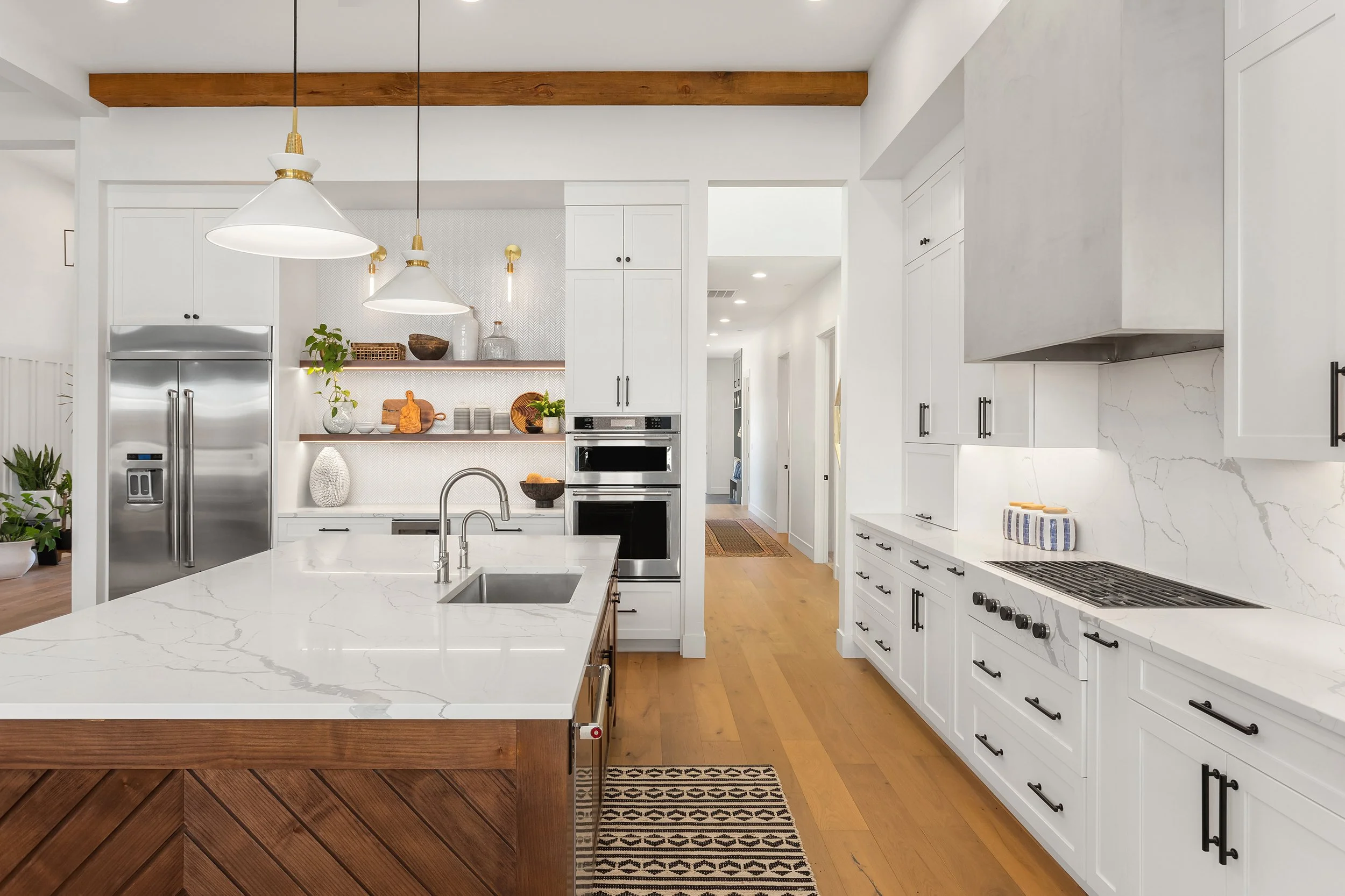 Modern kitchen with white cabinets, marble countertops, and stainless steel appliances. A central island with a white marble surface and wood accents. Pendant lights hanging from the ceiling, hardwood flooring, and decorative items on open shelves.