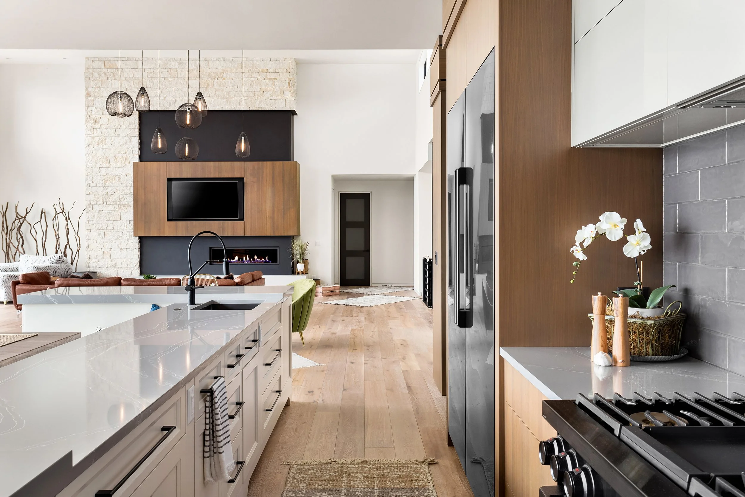 Modern kitchen with white cabinets, gray countertops, wooden accents, and a view of the living room with a fireplace and hanging pendant lights.