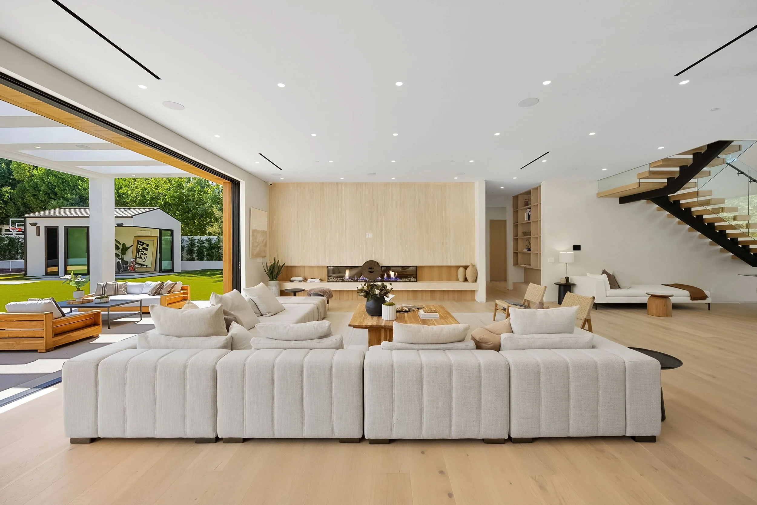 Spacious modern living room with large open sliding glass doors, view of backyard with small shed, and a sleek fireplace, wood flooring, and contemporary furniture.