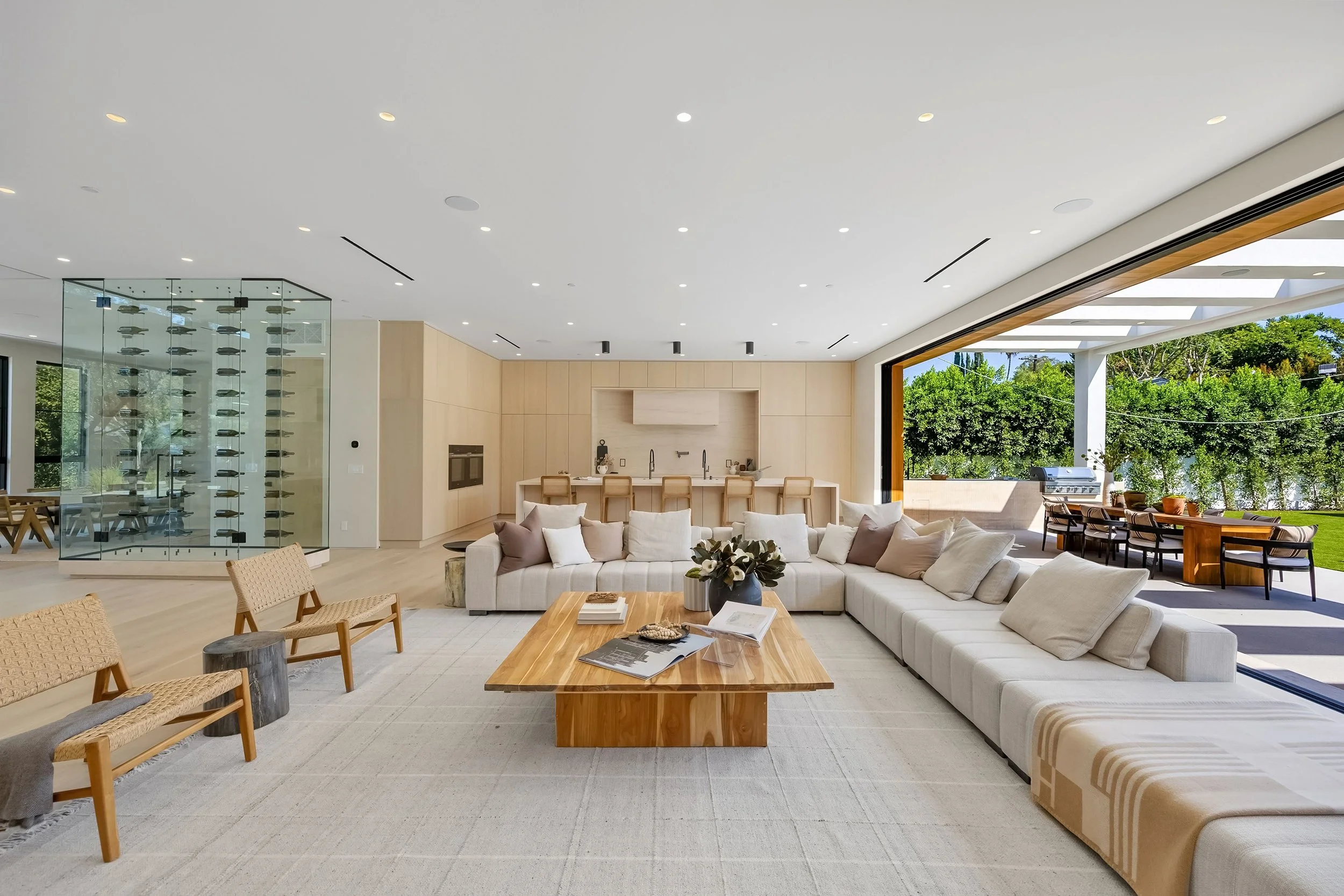 Open-concept living room with a large white sectional sofa, a wooden coffee table, and a modern kitchen with a bar area in the background. Large sliding glass doors reveal a lush green outdoor space.