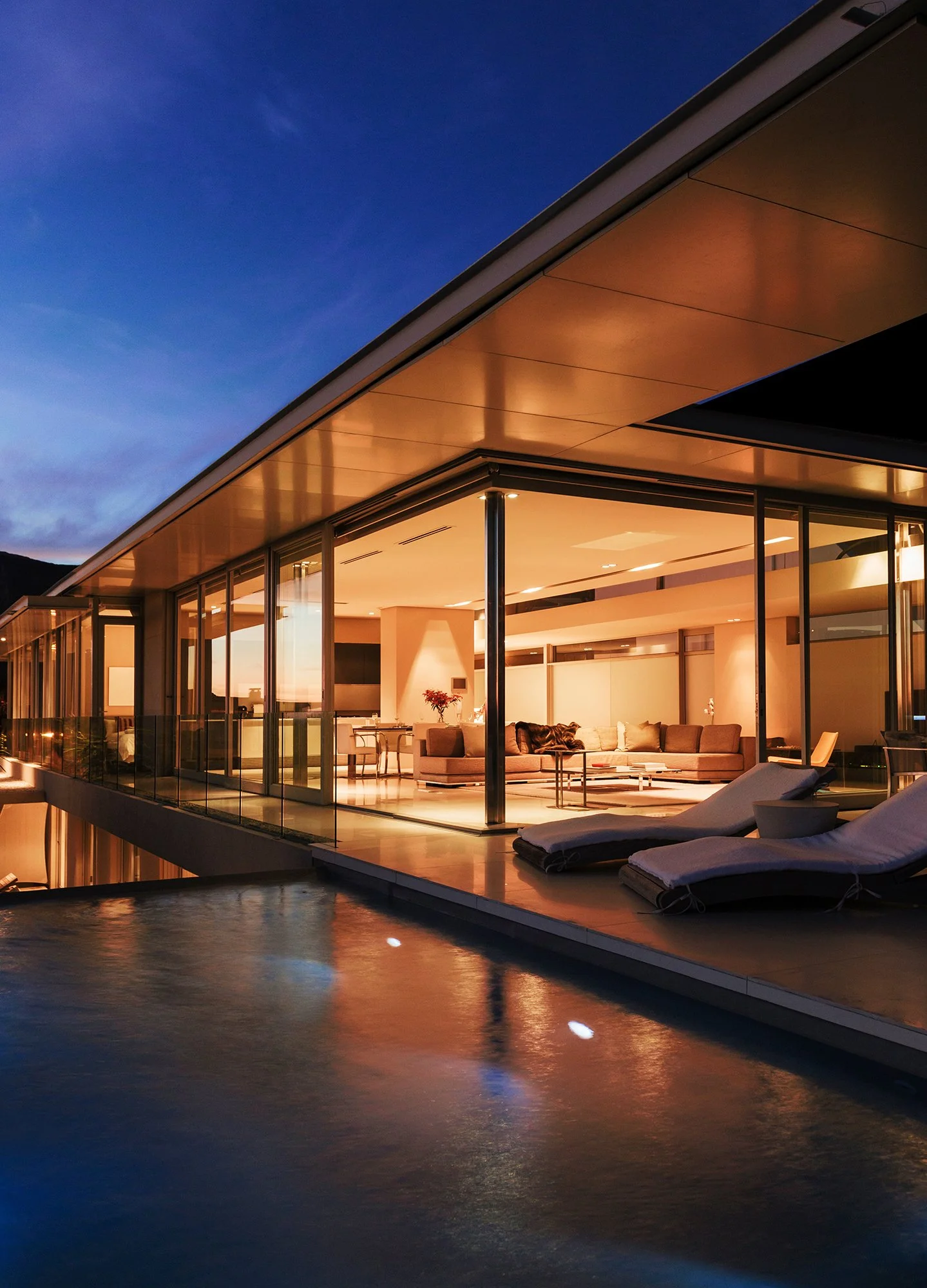 Modern house with large glass windows and a patio, illuminated from within, overlooking a calm body of water at dusk.
