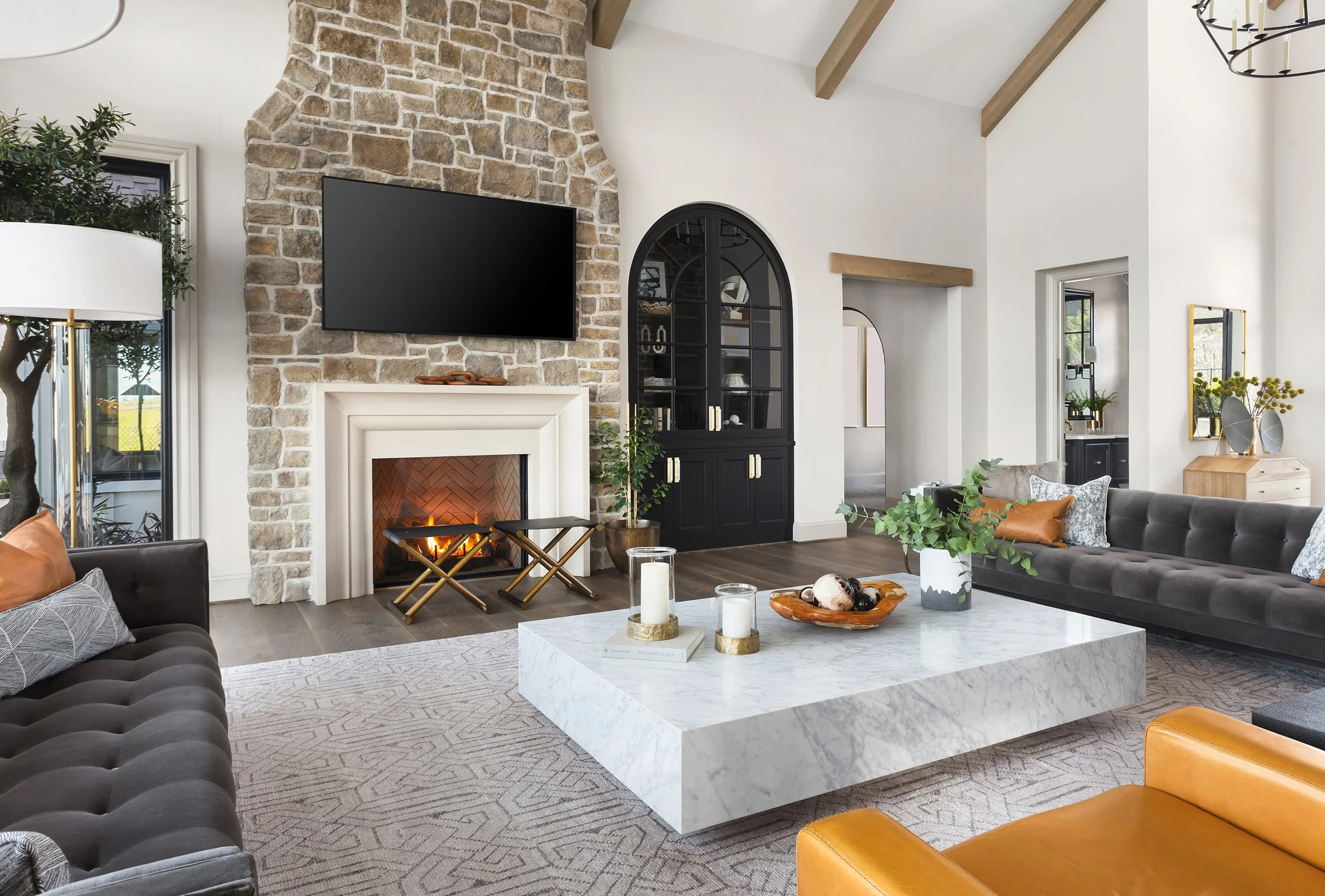 A spacious living room with a stone fireplace, a mounted flat-screen TV, a black cabinet, and a large marble coffee table surrounded by gray and orange furniture. Decor includes candles, books, potted plants, and artwork, with natural light coming through windows.