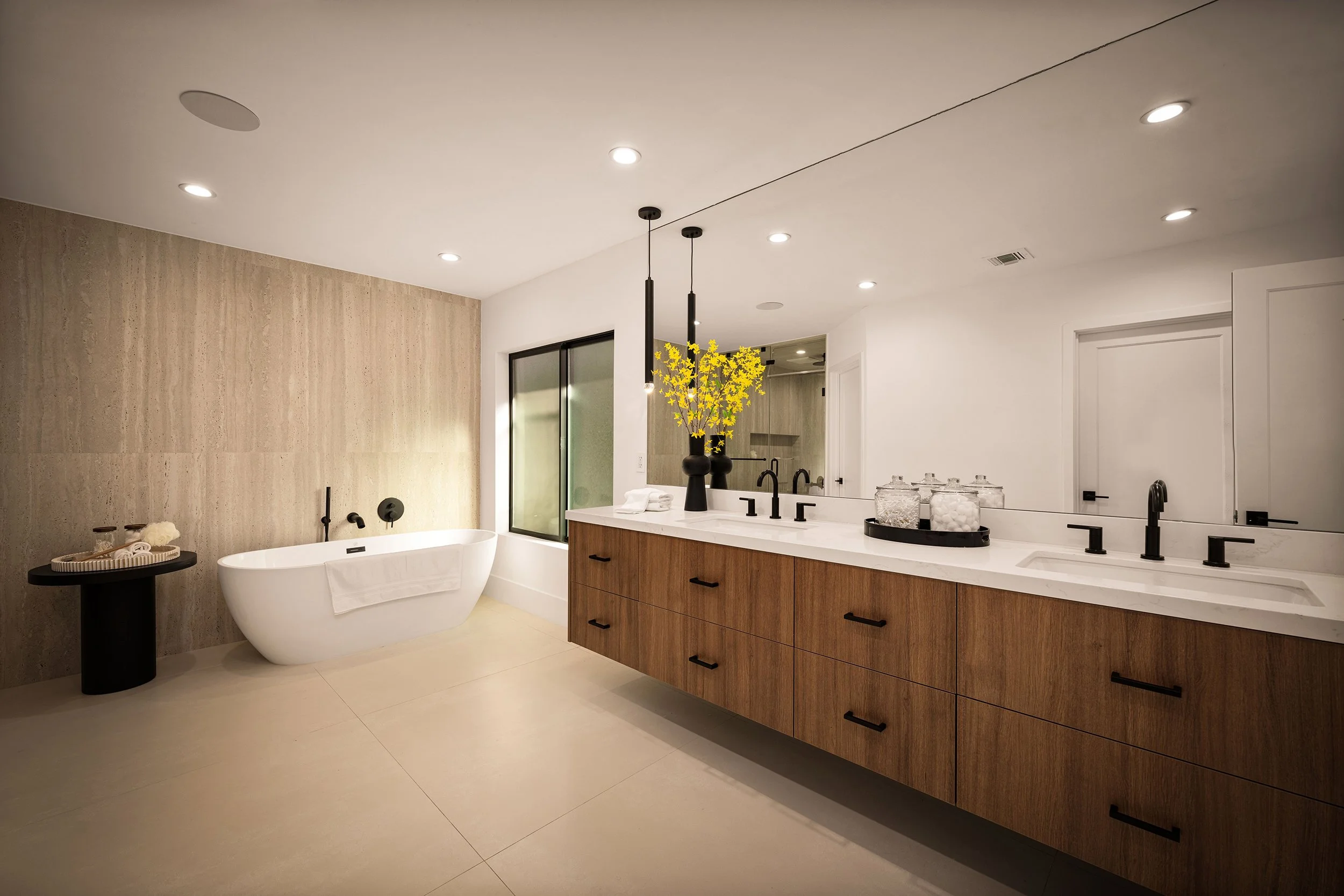 Modern bathroom with a freestanding bathtub, a double vanity with black fixtures, a large mirror, decorative glass jars, a yellow flowering plant, wooden cabinetry, and a beige textured accent wall.
