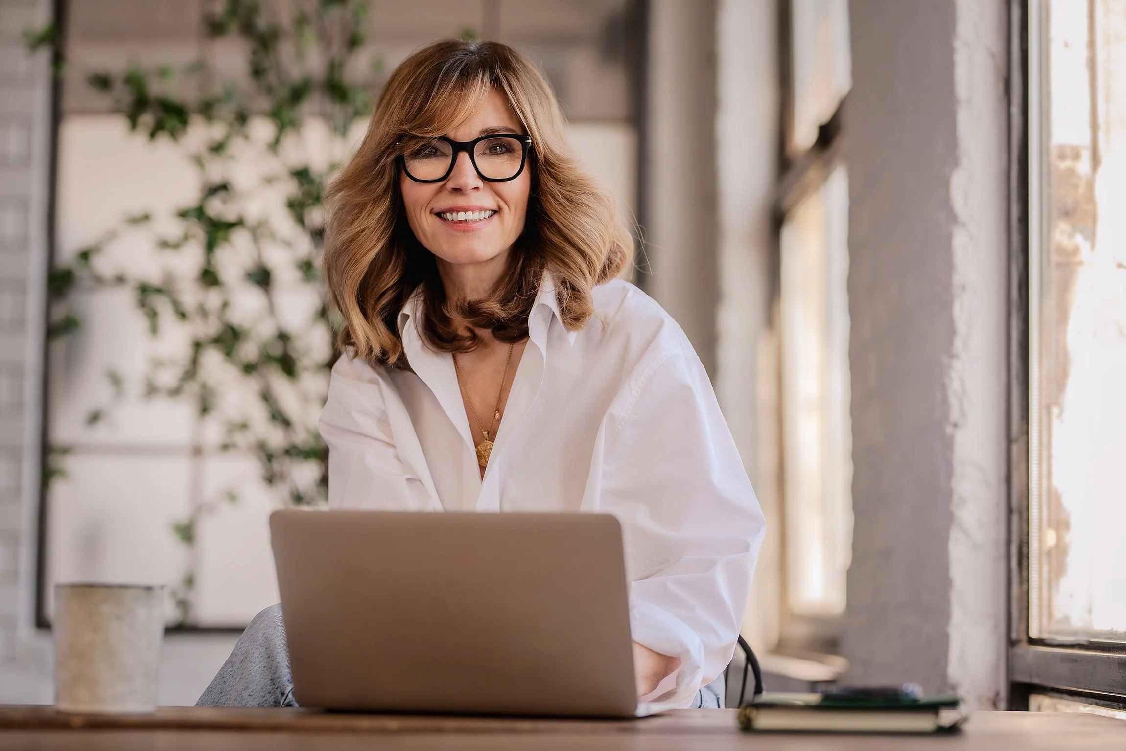 A woman with wavy, shoulder-length blonde hair and black-rimmed glasses smiling while sitting at a desk with a laptop in a bright room.
