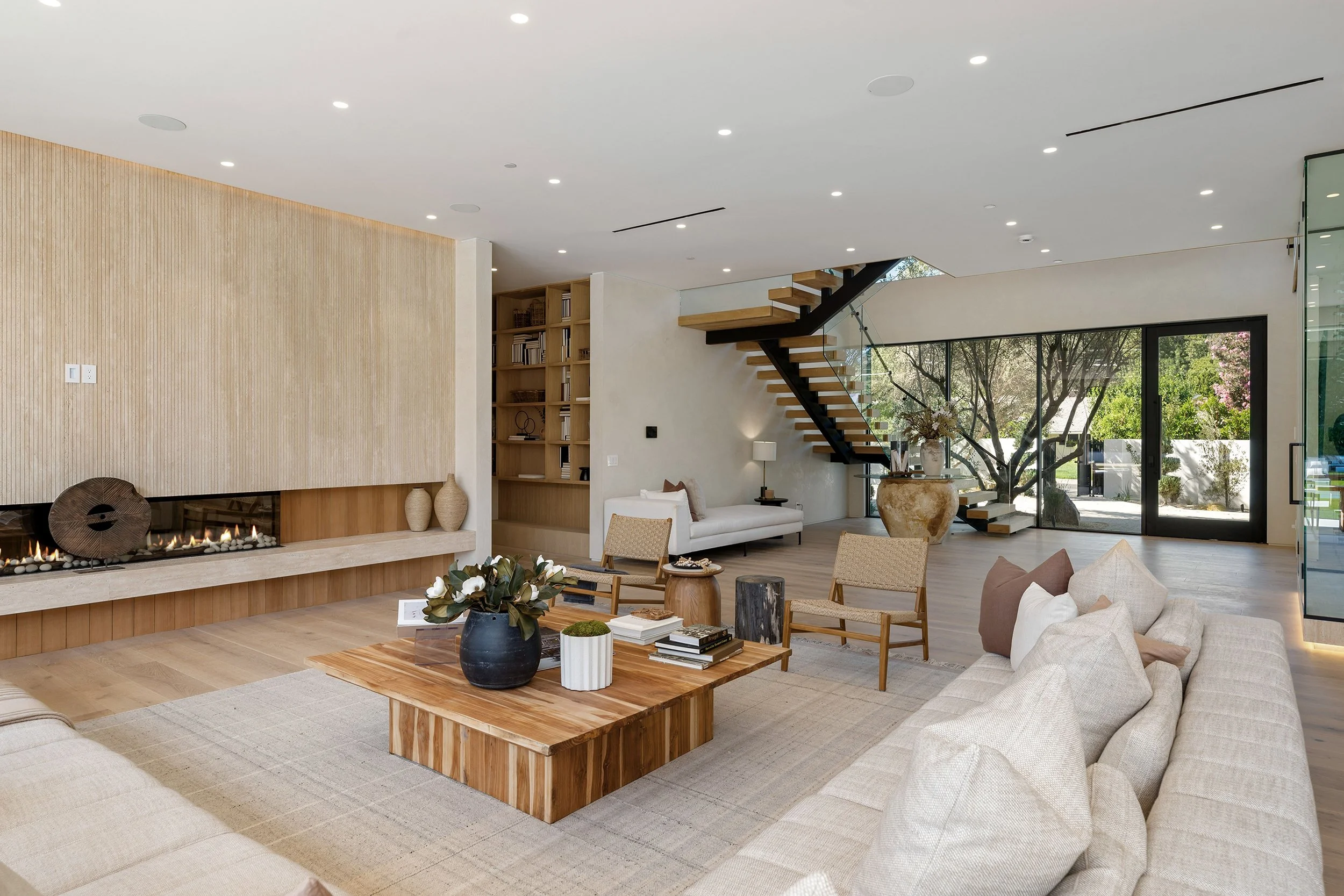 Modern living room with a neutral color palette, featuring a fireplace, a white sofa, wooden furniture, and large glass sliding doors showcasing an outside garden with trees and pink blossoms.