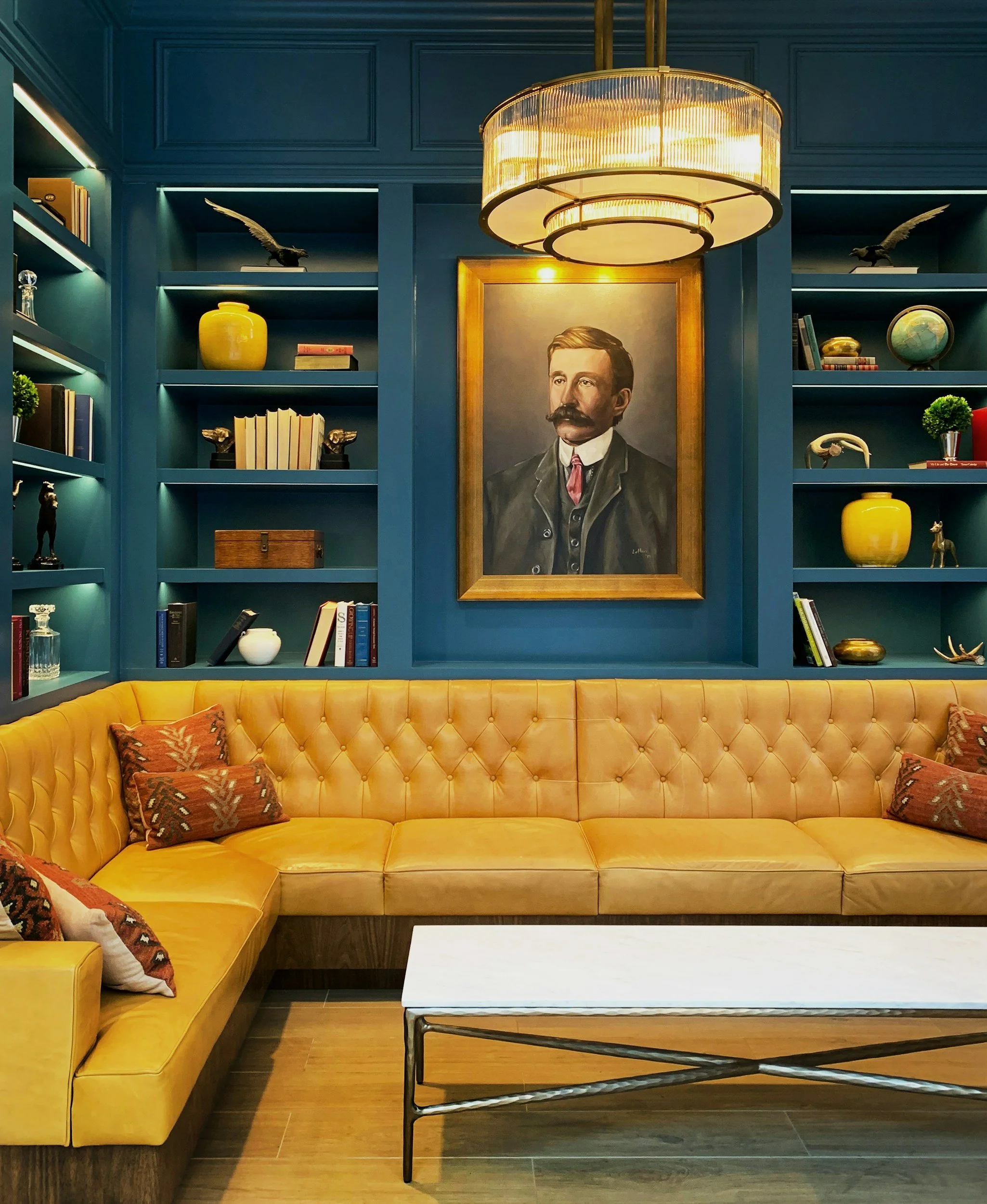 A yellow tufted sofa with decorative pillows, a white marble coffee table, a blue bookshelf with decor items including a bird sculpture, a yellow vase, books, and a globe, a framed painting of a man with a mustache, and a large circular chandelier with warm lighting.