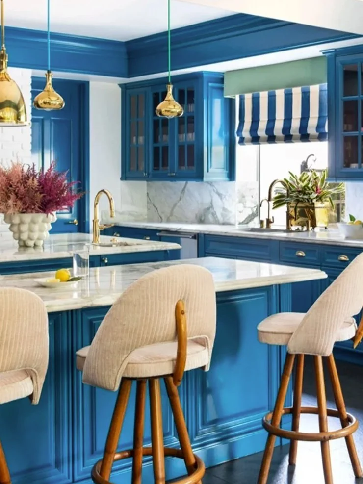 Colorful kitchen with blue cabinets, white marble countertop, gold fixtures, beige upholstered bar stools, potted plants, and a window with a striped blue and green valance.