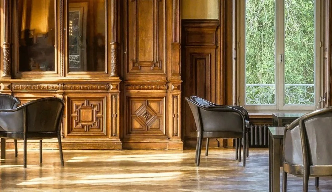 A room with ornate wood paneling and large window with a view of green trees outside, several black chairs around a small black table, and a wooden floor.
