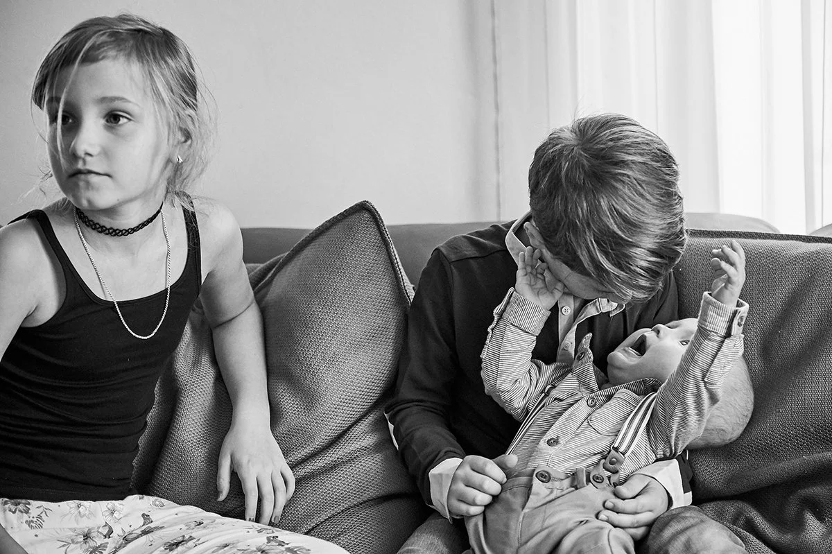Drei Kinder, ein Mädchen und zwei Jungen, sitzen auf einem Sofa. Das Mädchen steht rechts, der ältere Junge sitzt in der Mitte, hält den kleinen Jungen im Arm, der ausdrucksstark in die Kamera schreit, während der ältere Junge ihm gegen das Ohr greif