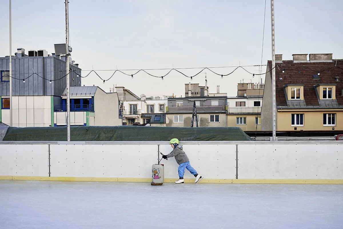 Ein Kind in Eishockeykleidung, das auf einer Eisbahn mit einem Koffer mit Schneemann-Aufdruck übt, während im Hintergrund eine Stadt mit Gebäuden zu sehen ist.