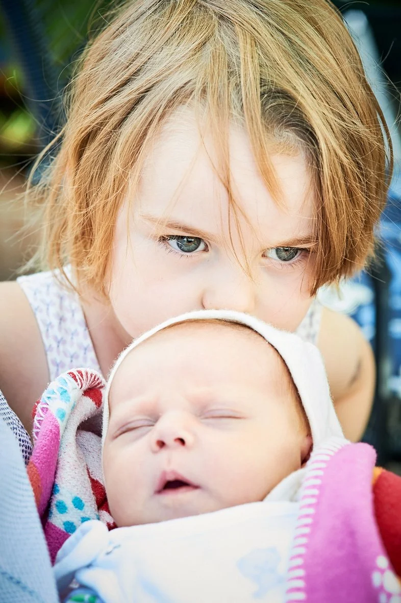Ein kleines Kind schaut aufmerksam auf ein schlafendes Baby, das in einem weißen Hut und einem bunten Handtuch eingewickelt ist.