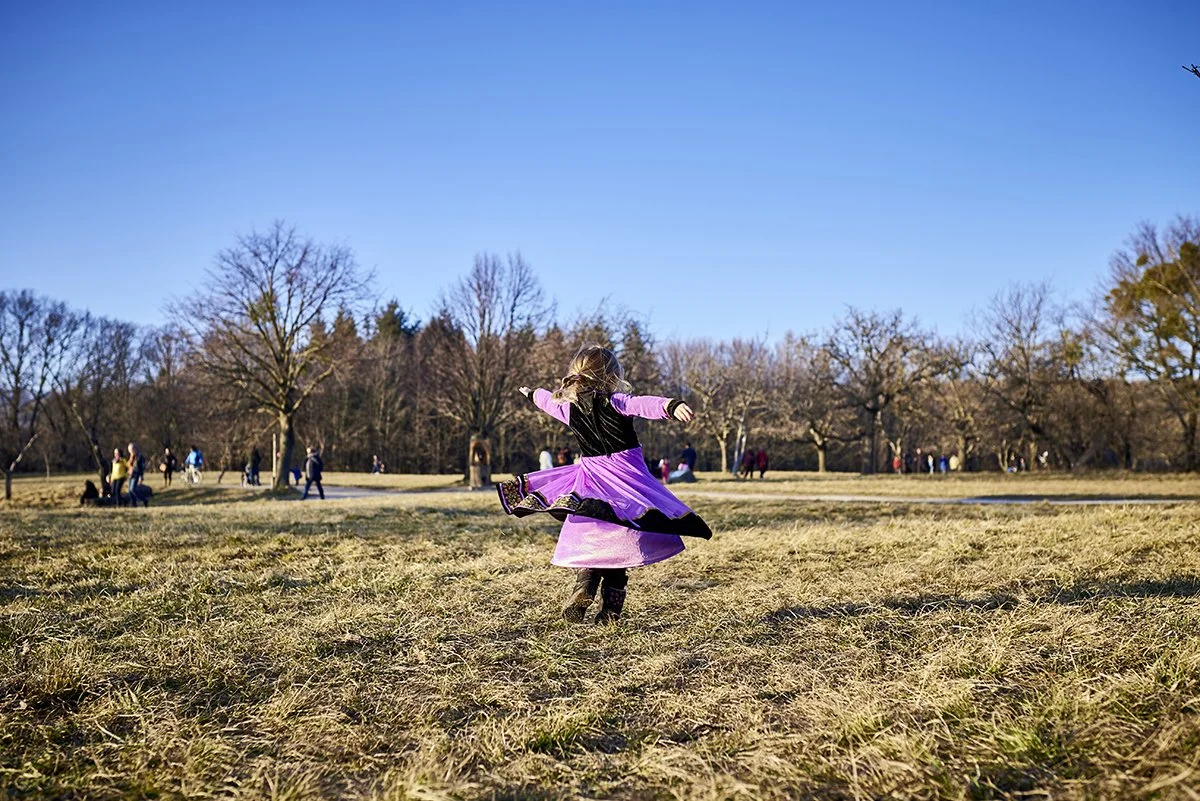 Ein junges Mädchen in einem pinken Kleid tanzt auf einer Wiese in einem Park bei sonnigem Wetter, im Hintergrund sind Bäume und mehrere Menschen zu sehen.