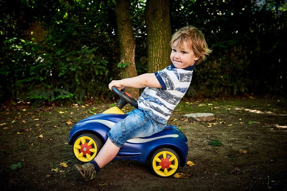 Ein junger Junge sitzt auf einem Spielzeugauto im Park, umgeben von Bäumen und Laub auf dem Boden.