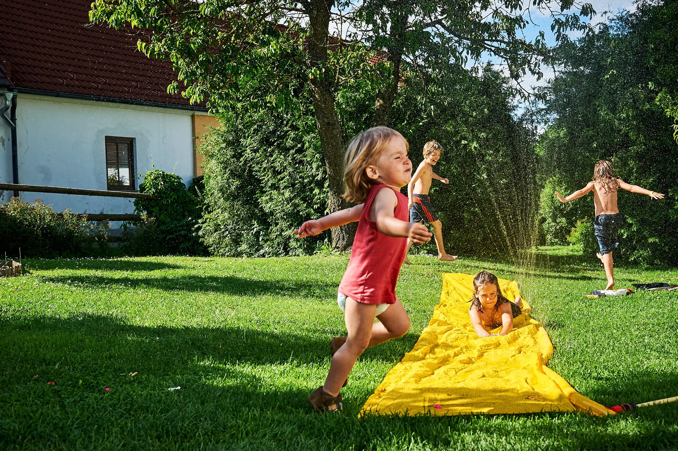 Kinder spielen im Garten mit einem Wasserbalken und Wasser spritzt in die Luft, während die Sonne scheint.
