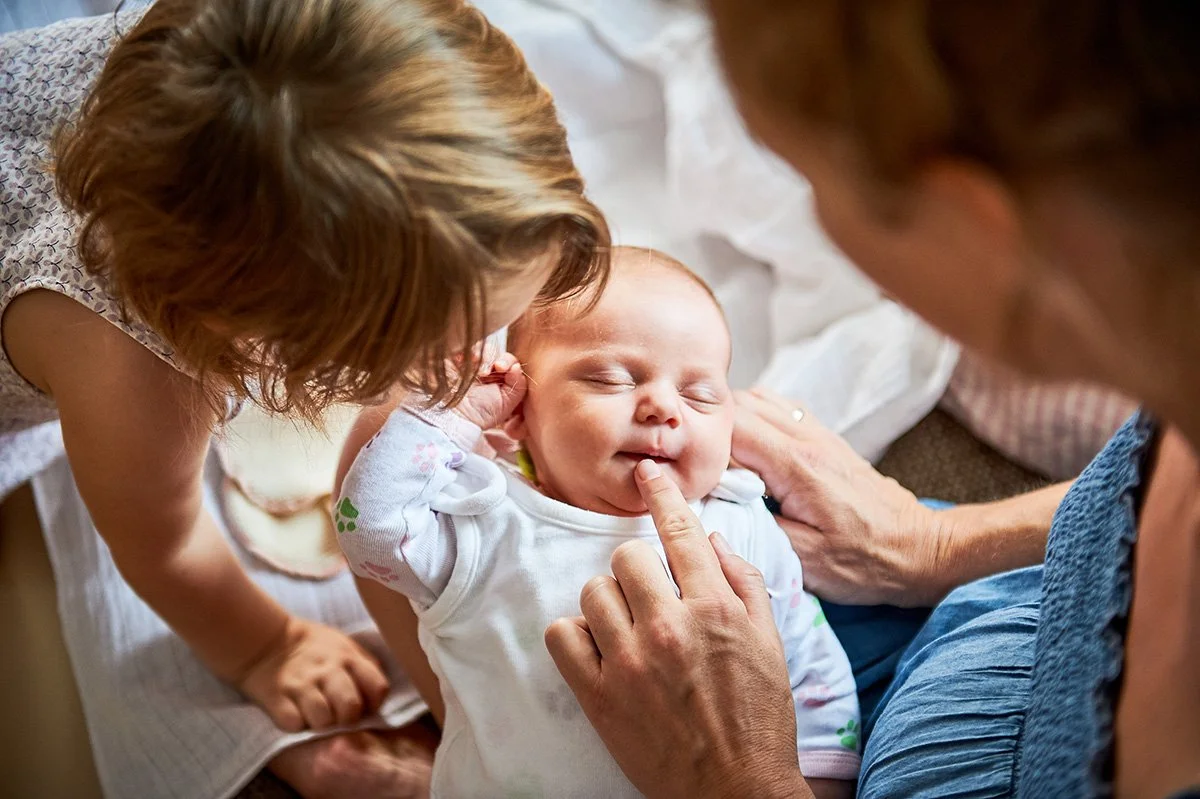 Ein Baby liegt glücklich auf einem Bett, während eine Frau, möglicherweise die Großmutter, es liebevoll streichelt. Eine andere Frau, vermutlich die Mutter, ist neben ihnen und zeigt auf die Wange des Babys, während sie lächeln.