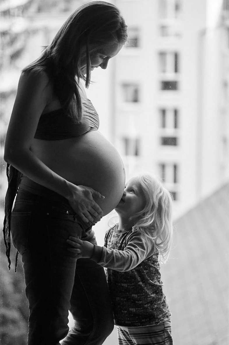 Eine schwangere Frau mit ihrer kleinen Tochter, die an ihrem Bauch schnüffelt, vor einem Fenster in einem Innenraum.