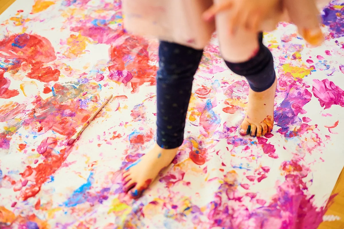 Ein Kind, das auf einem weißen Papier mit Fingerfarben malt, umgeben von bunten Farbklecksen und -spuren.