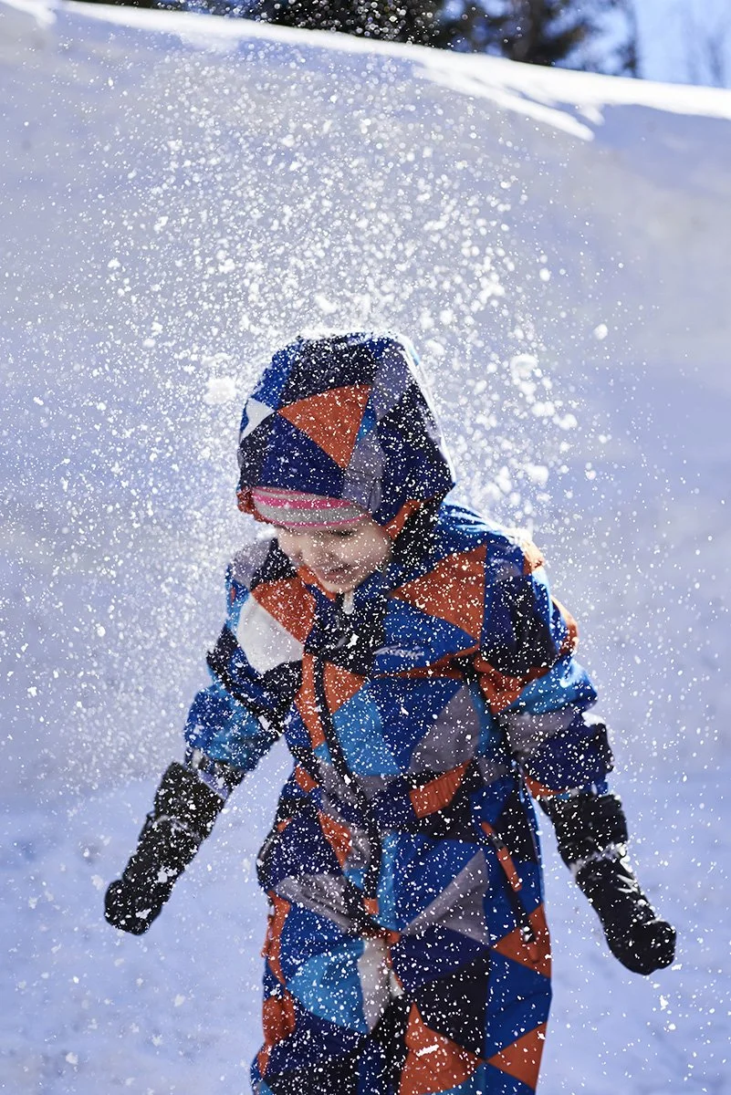 Ein Kind in bunter Winterkleidung spielt im Schnee, während Schneeflocken um es herumwirbeln.