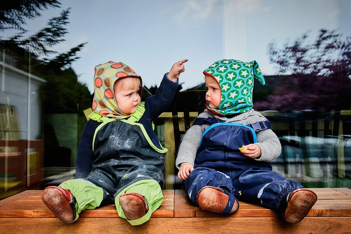 Zwei kleine Kinder sitzen draußen auf einer Holzbank, tragen Regenkleidung und Mützen, und scheinen ein Gespräch zu führen.