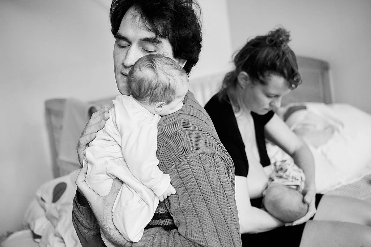 Ein Mann hält ein Baby, während eine Frau ein Baby im Arm hält und auf einem Bett sitzt.