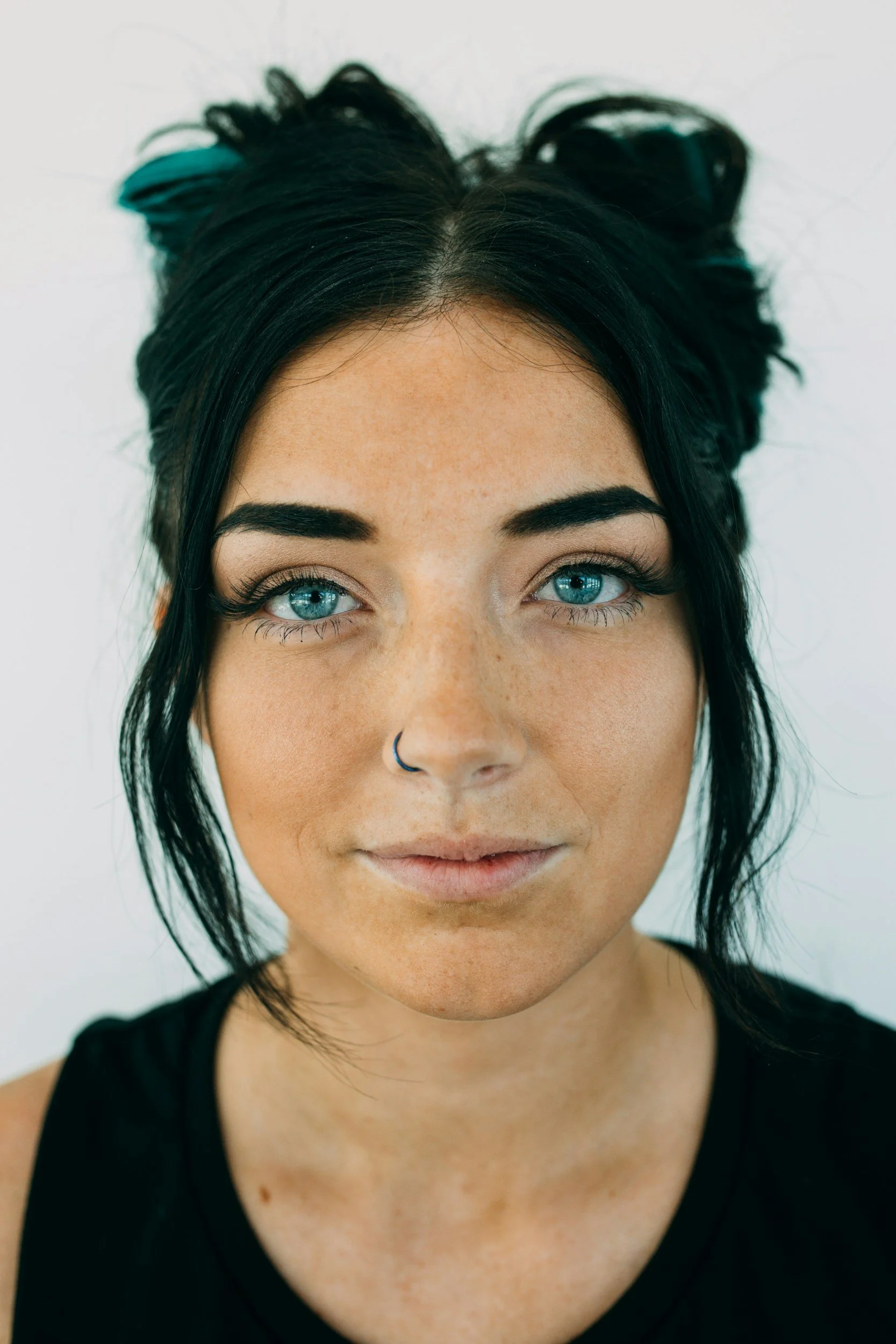 Close-up of a woman with black hair styled in messy buns, bright blue eyes, and a nose ring, wearing a black top, against a plain white background.