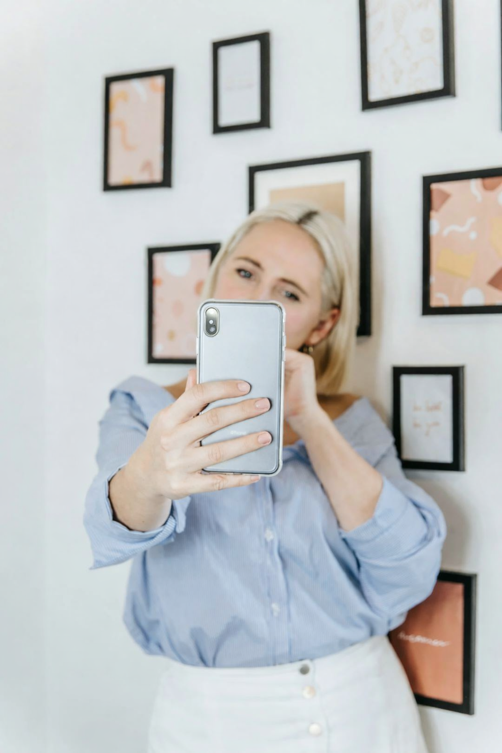 A woman with blonde hair taking a selfie with her smartphone in a room with abstract art on the wall.
