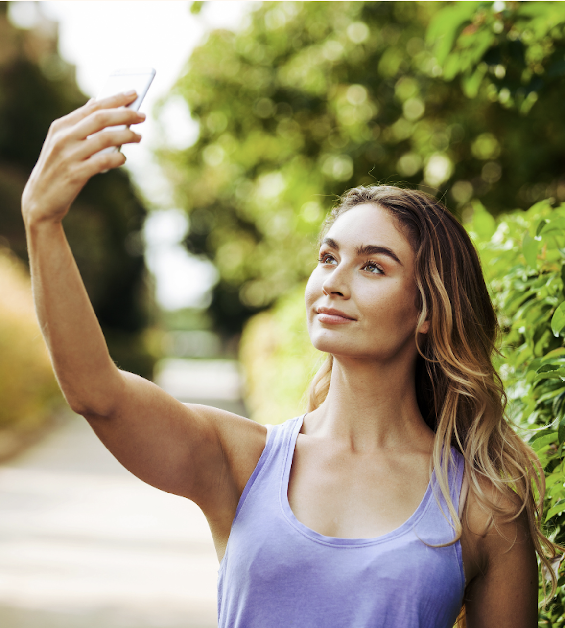 A woman with long, wavy hair taking a selfie outdoors with trees and greenery in the background