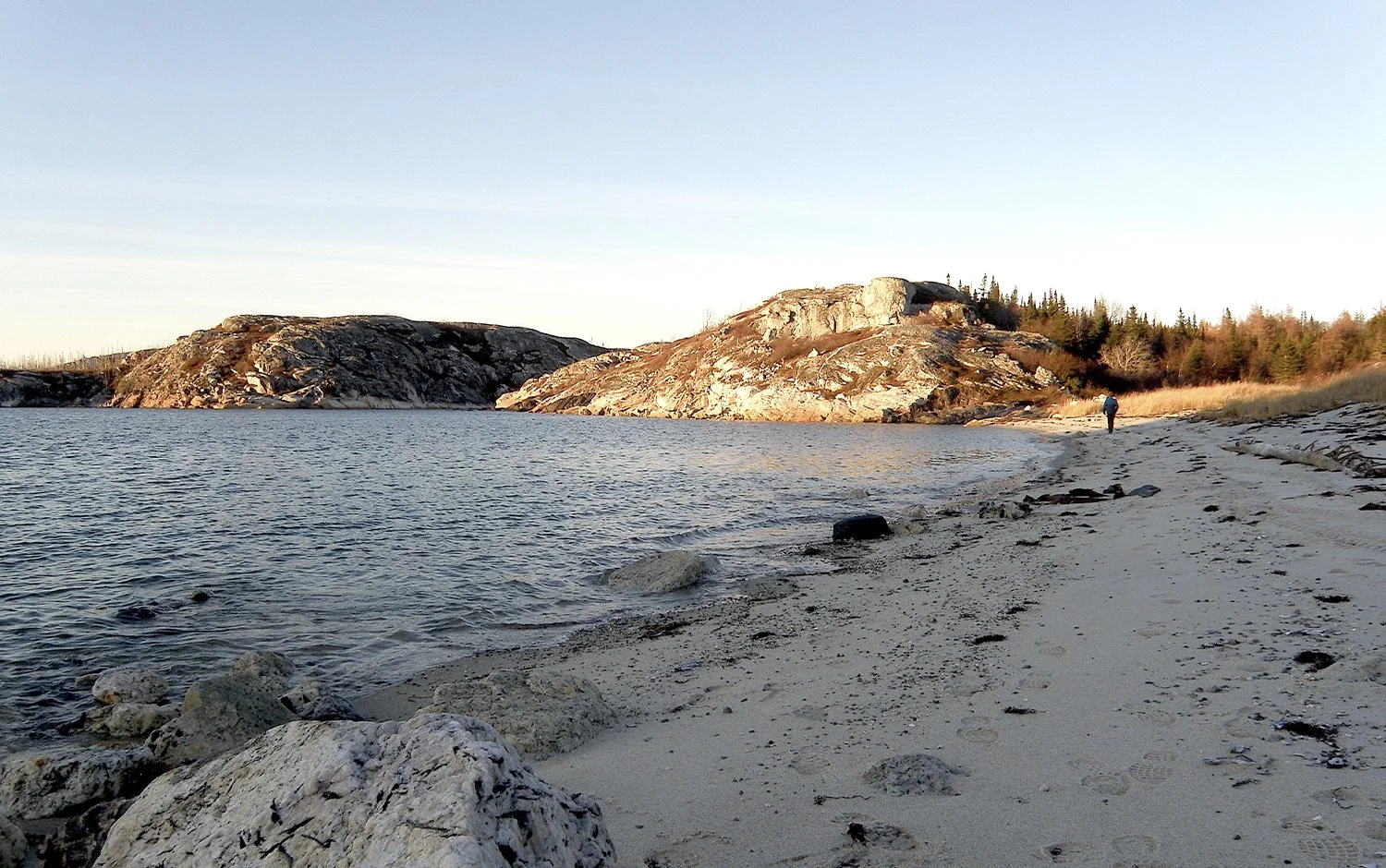 Une plage rocheuse et peu peuplée avec un seul individu marchant sur le sable, des rochers au premier plan, une étendue d'eau calme, des collines rocheuses en arrière-plan et un ciel dégagé, dans une atmosphère paisible.
