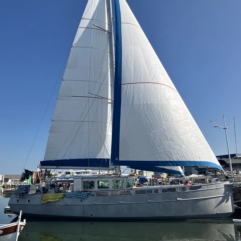 Voilier à voile blanche amarré dans un port, avec des voiles déployées et une cabine visible, fond bleu clair.