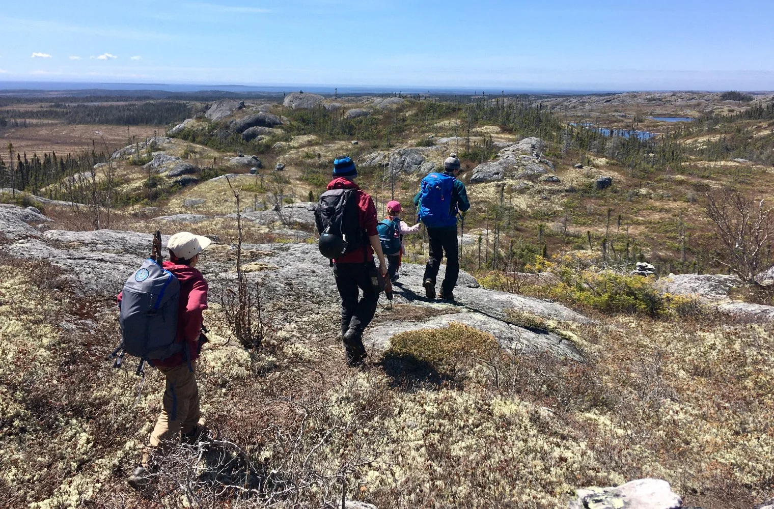 Groupe de personnes faisant de la randonnée en montagne sous un ciel clair, avec des rochers et une végétation sparse en premier plan et des collines et plans d'eau à l'horizon.
