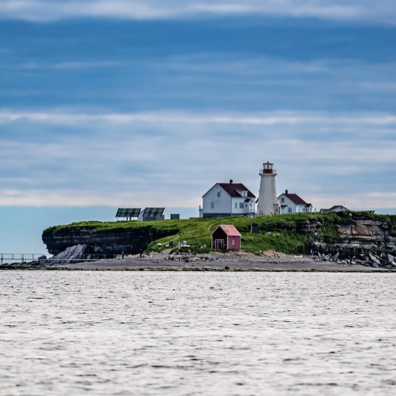 Phare blanc avec maison adjacente sur une falaise herbeuse, petit bâtiment rouge en premier plan, panneaux solaires, mer au premier plan, ciel nuageux.