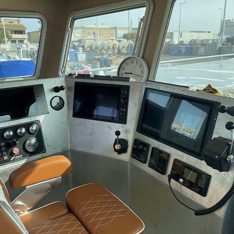 Intérieur d'une cabine de contrôle avec plusieurs écrans, un compteur, un joystick, une montre, et une chaise en cuir marron, devant de grandes fenêtres donnant sur un port ou une zone industrielle.