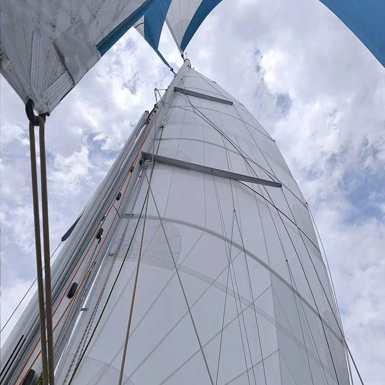 Photo prise sous un mât de catamaran montrant la voile principale, les cordages et le mât avec un ciel nuageux en arrière-plan.