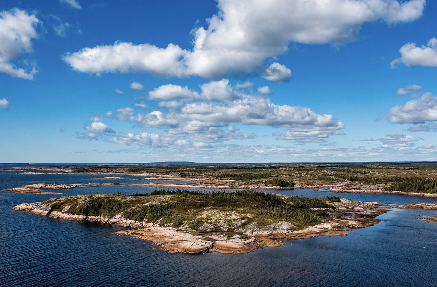 Une vue aérienne d'une côte rocheuse avec des eaux bleues, sous un ciel bleu parsemé de nuages blancs.