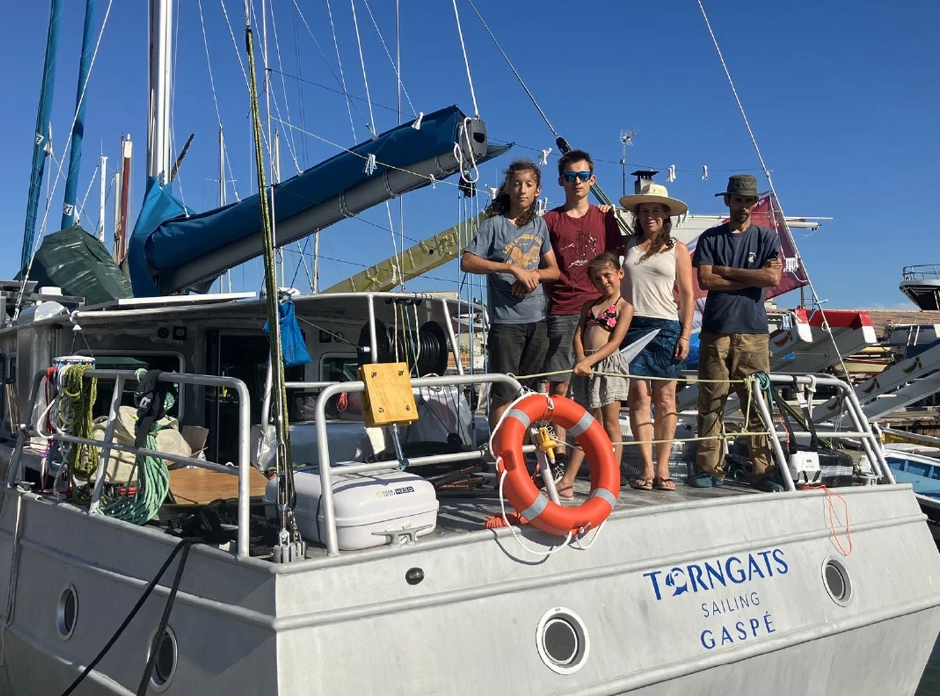 Groupe de six personnes sur un bateau à voile nommé 'TORNGATS' au port, avec des voiliers en arrière-plan, sous un ciel bleu