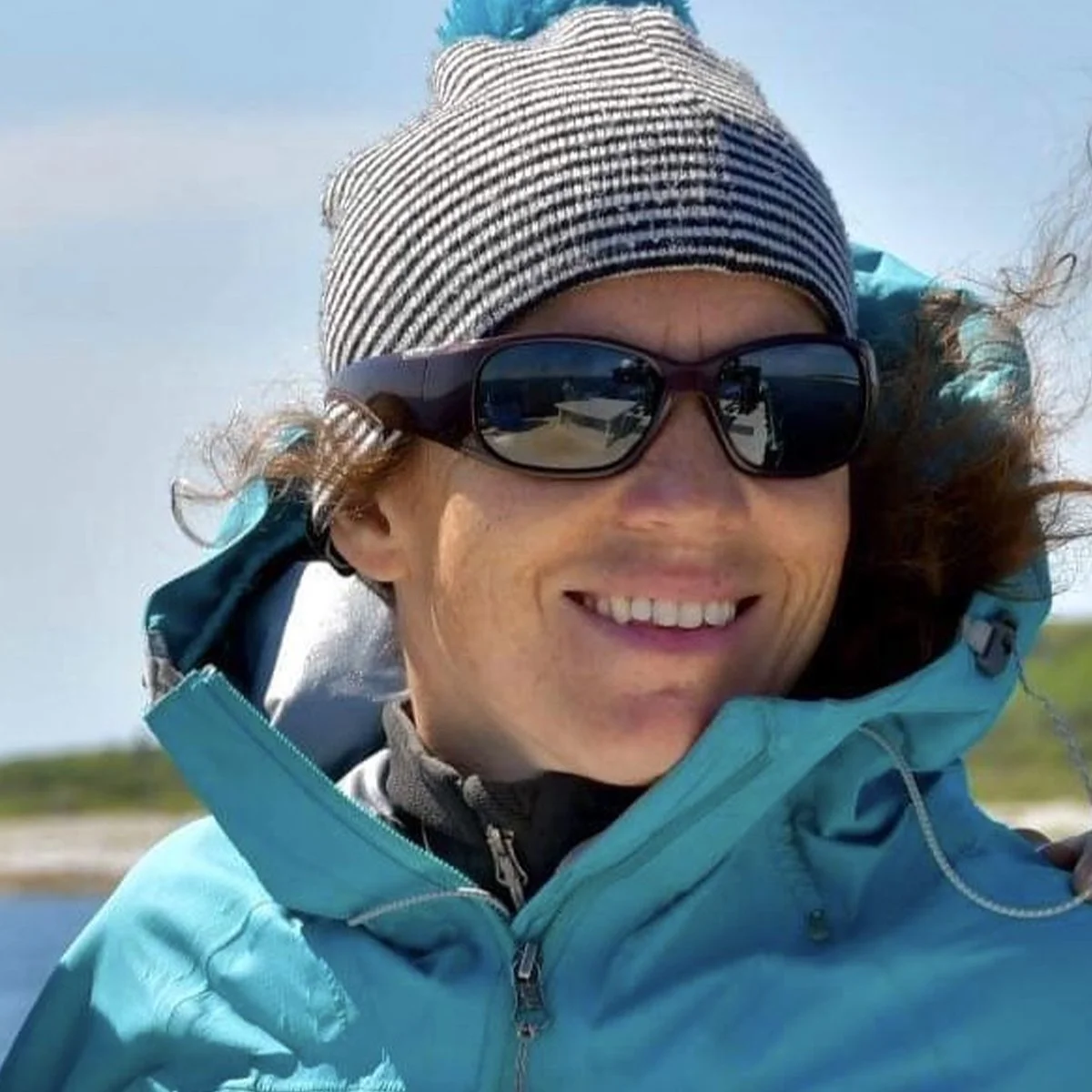 Femme souriante portant une veste bleue, des lunettes de soleil noires et un bonnet rayé, à l'extérieur par temps ensoleillé.