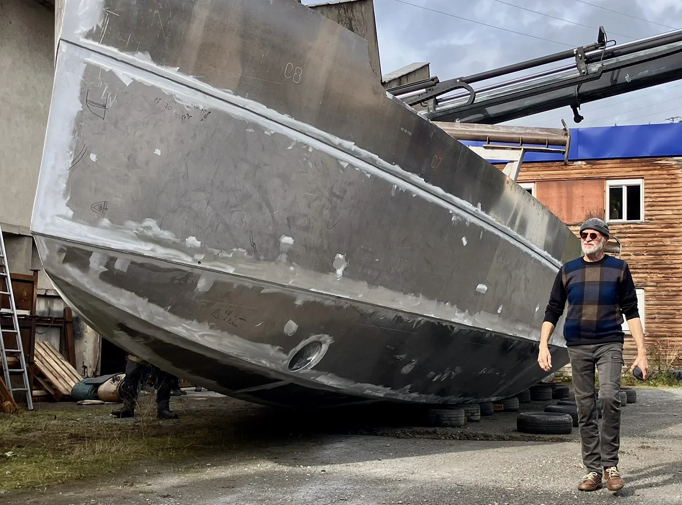 Un homme portant des lunettes de soleil marche à côté d'un grand bateau en construction en acier, placé sur des pneus, dans une zone de travail en extérieur.