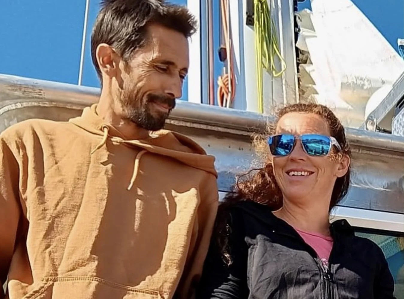 Un homme et une femme souriants, assis côte à côte, avec un équipement d'escalade en arrière-plan, dans un environnement en plein air, sous un ciel bleu.