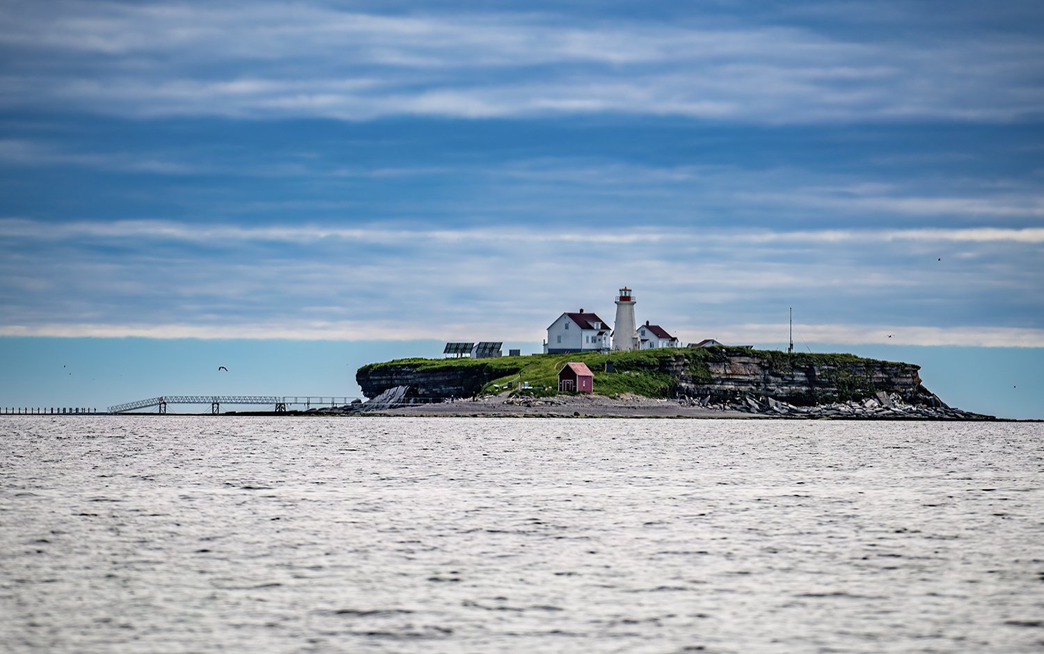 Île avec un phare blanc, maisons, et un hangar rouge, entourée d'eau et un ciel nuageux.