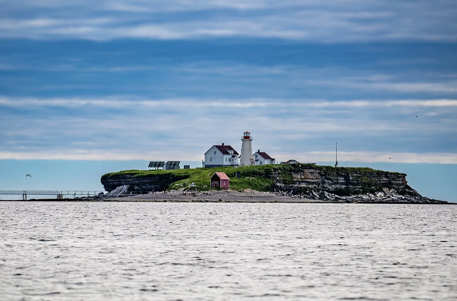 Phare sur une île rocheuse avec bâtiments, sous un ciel nuageux, vue à partir de la mer.
