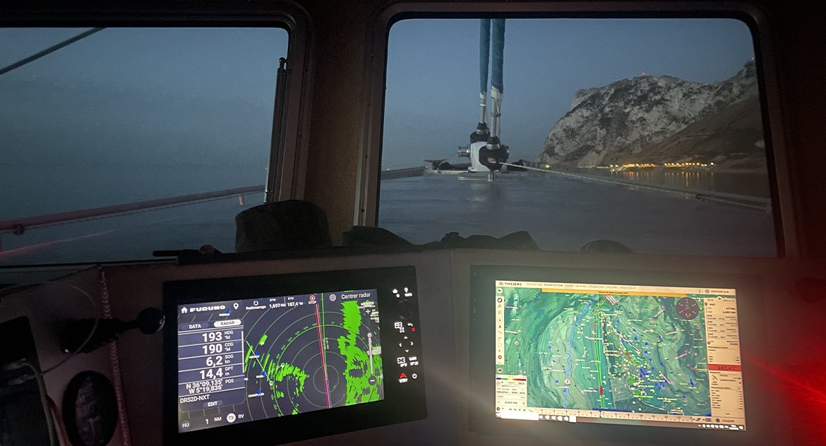 Vue de l'intérieur d'un bateau avec deux écrans de navigation, une mer calme, une montagne en arrière-plan et le ciel au crépuscule.