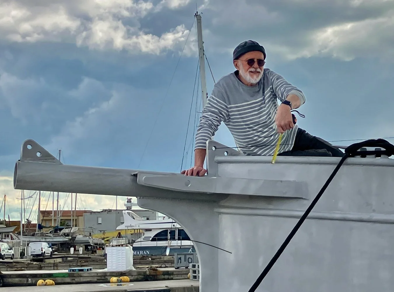 Un homme âgé souriant, portant un bonnet et des lunettes de soleil, est assis sur une embarcation au port, avec un ciel nuageux en arrière-plan.