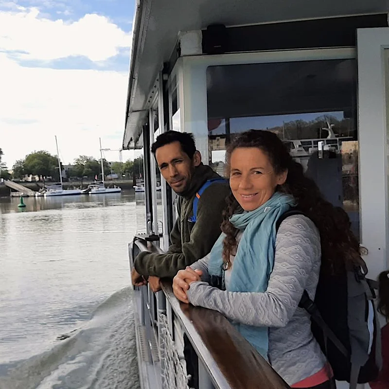 Deux personnes regardent par la fenêtre d'un bateau alors qu'il navigate sur l'eau, avec des voiliers et des arbres en arrière-plan.