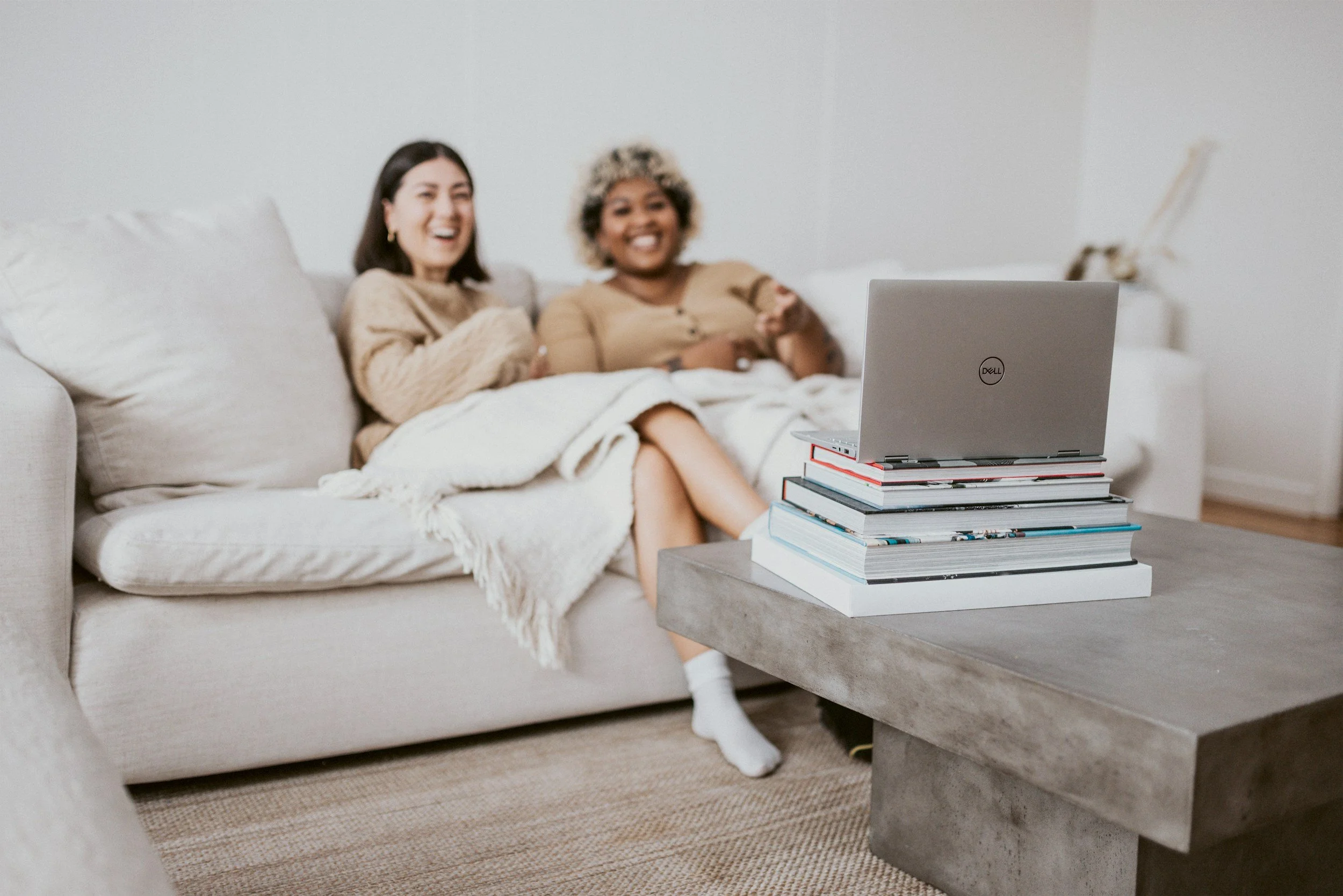 Two women sitting on a beige couch, watching a laptop display. They appear to be happy and laughing. The couch has a light blanket draped over it. In front of them, on a coffee table, there is a stacked arrangement of books with a gray 'Dell' laptop on top. The room has a neutral color scheme with white walls and a beige rug.