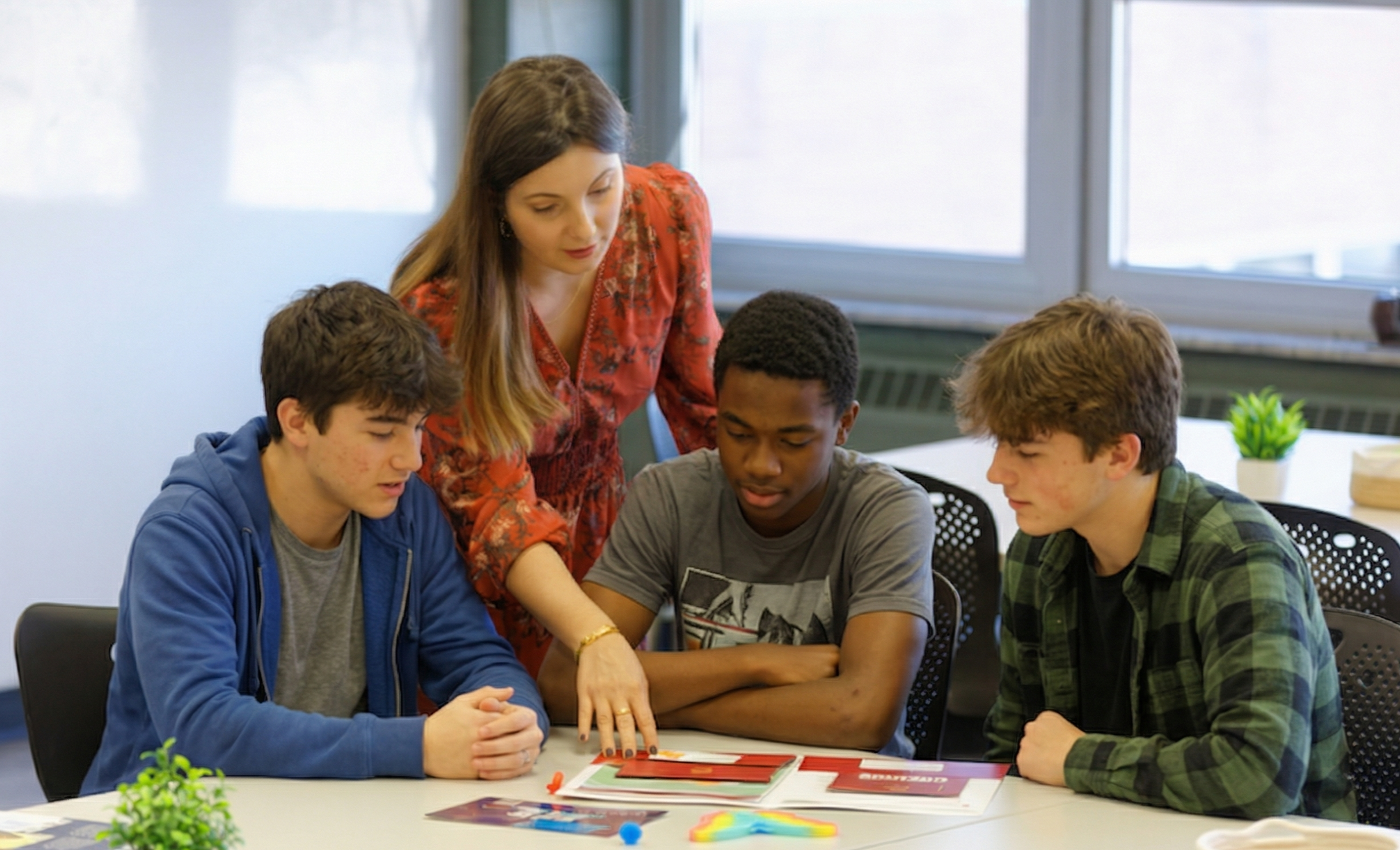 A teacher guides a group of four male students sitting around a table, looking at materials in a classroom with large windows.