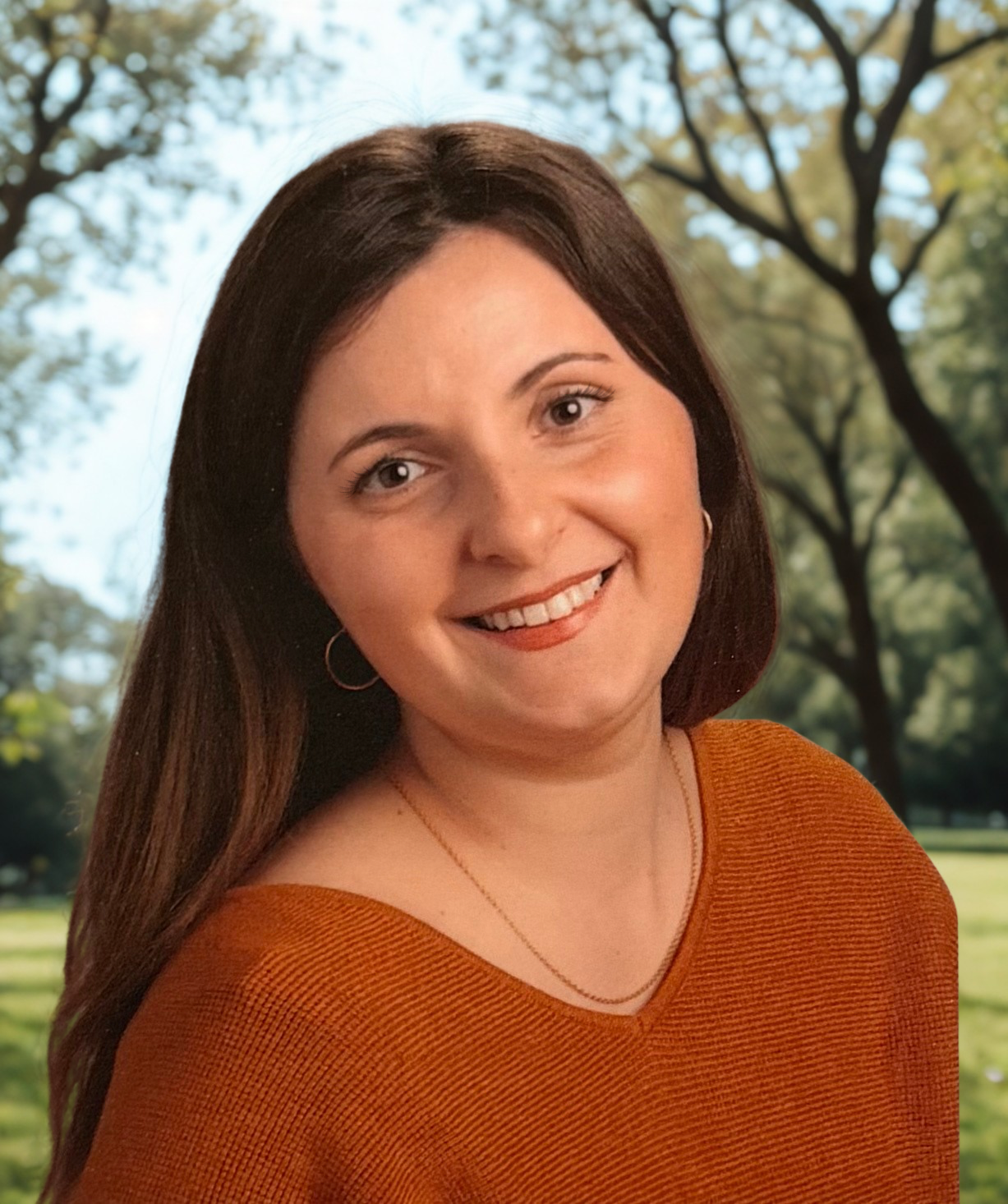 Smiling woman with brown hair, wearing an orange top, posed outdoors with trees and sky in the background.