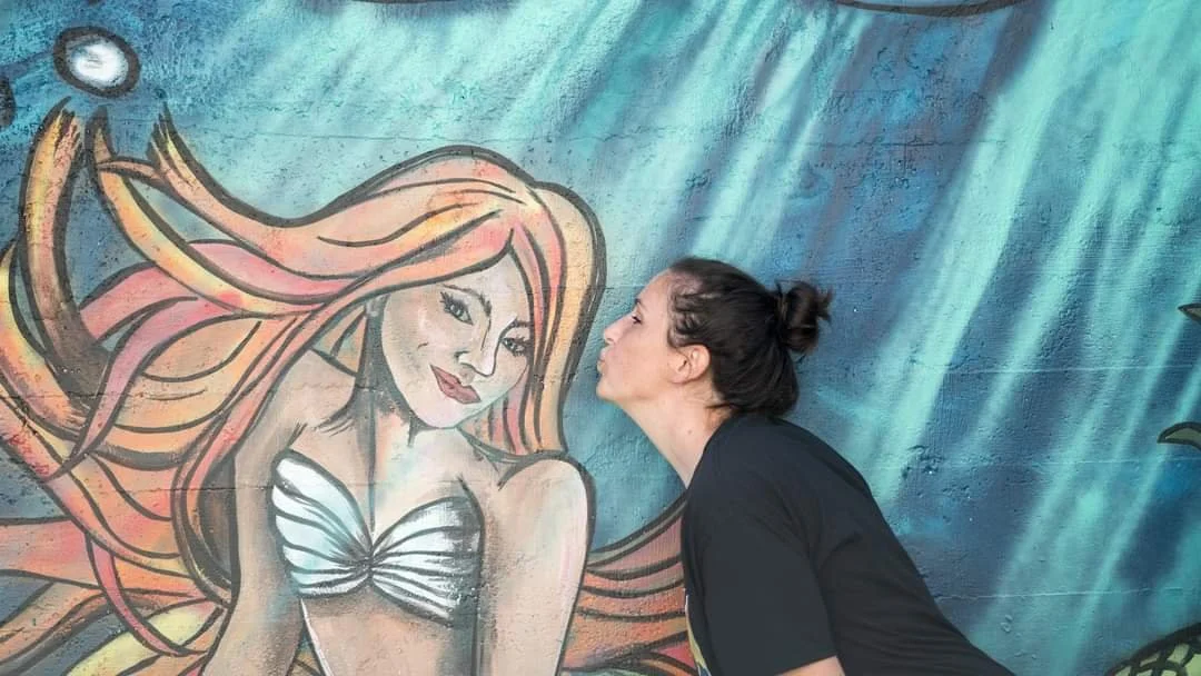 A woman is kissing a large mural of a mermaid painted on a wall.