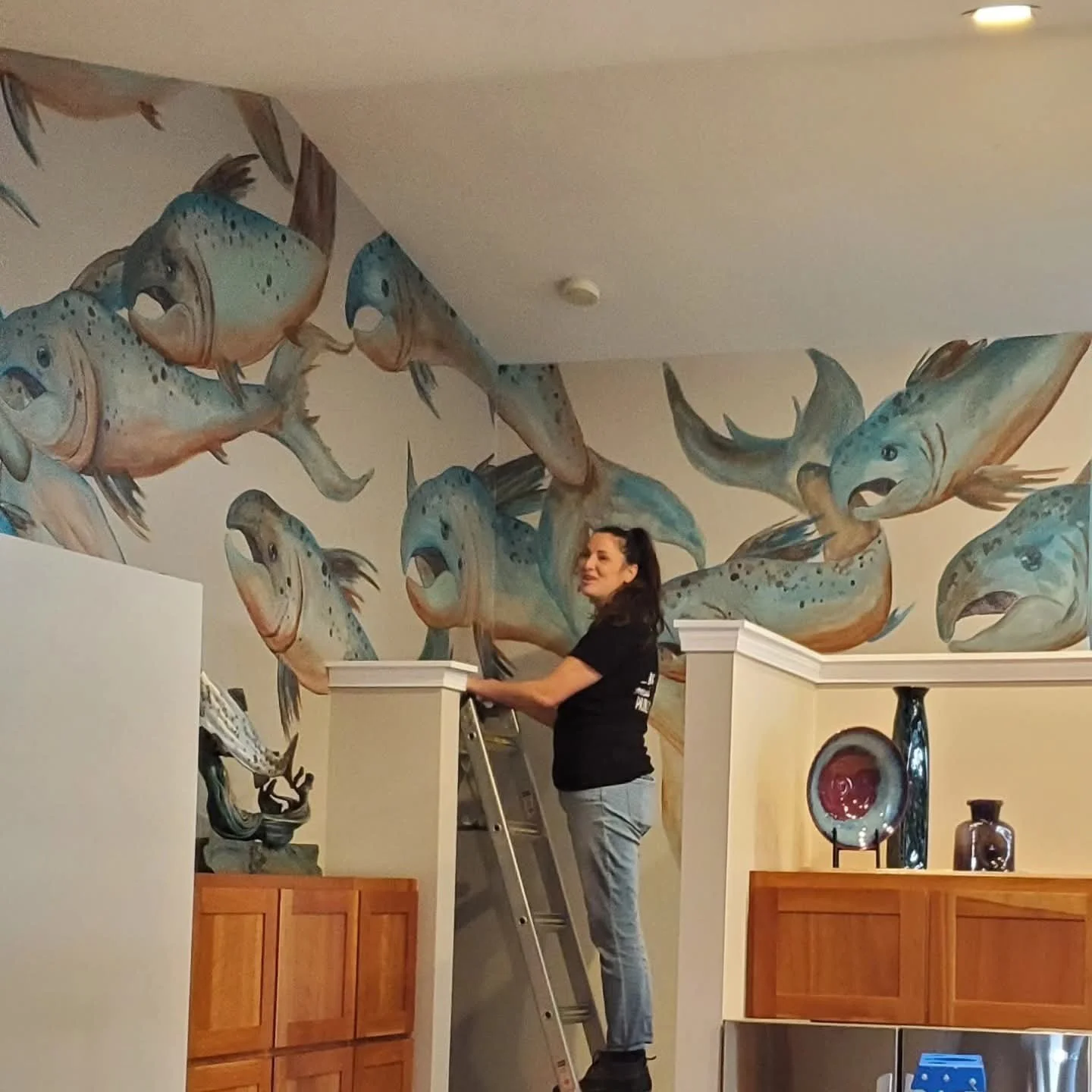 A woman standing on a ladder decorating a wall with a mural of large, colorful fish in a room with wooden cabinets and decorative objects.